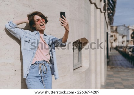 Young smiling caucasian excited fun woman 20s wear jeans clothes glasses do selfie shot on mobile phone post photo on social network outdoors near city street building wall People lifestyle concept.