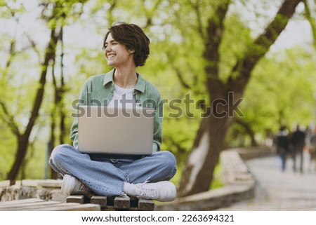 Full length young fun happy student freelancer woman 20s in green jacket jeans sit on bench in spring park outdoors rest use laptop pc computer work online look aside People urban lifestyle concept