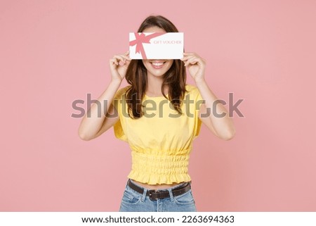 Smiing cheerful funny young brunette woman 20s wearing yellow casual t-shirt posing standing covering eyes with gift certificate hiding isolated on pastel pink color wall background studio portrait Royalty-Free Stock Photo #2263694363