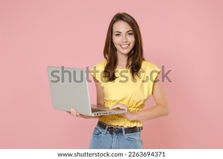 Smiling beautiful young brunette woman 20s wearing yellow casual t-shirt posing standing working on laptop pc computer looking camera isolated on pastel pink color wall background studio portrait