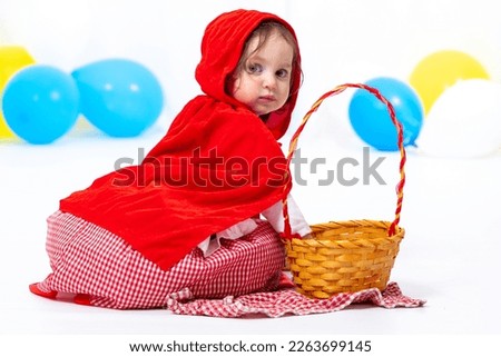 A little girl dressed as a red riding hood sits near a basket. Carnival, Purim, Halloween