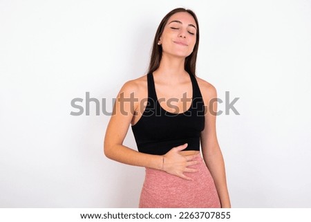 Satisfied smiling young woman wearing sportswear over white studio background , keeps hands on belly, being in good mood after eating delicious supper, demonstrates she is full. Pleasant feeling in st Royalty-Free Stock Photo #2263707855