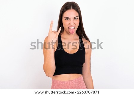 Portrait of a crazy young woman wearing sportswear over white studio background showing tongue horns up gesture, expressing excitement of being on concert of band.