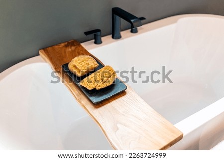 Bathtub wooden shelf caddy holder in bathroom staging model home house with sponge loofah scrubber in modern luxury white room Royalty-Free Stock Photo #2263724999