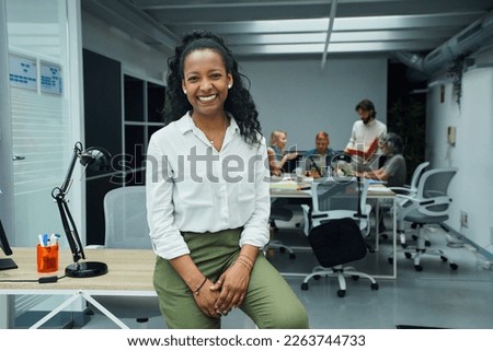 Portrait of smiling young African American professional female sitting at modern office desk. Happy mixed race business woman team leader posing for photo at corporate meeting. Concept of workplace.  Royalty-Free Stock Photo #2263744733