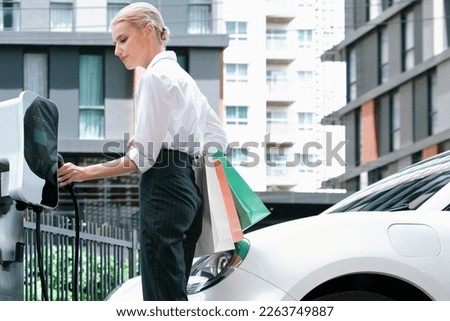 Recharging electric vehicle at charging station with businesswoman carrying shopping bag. Progressive lifestyle of a man in city with ecological concern for clean electric energy driven car ideal.