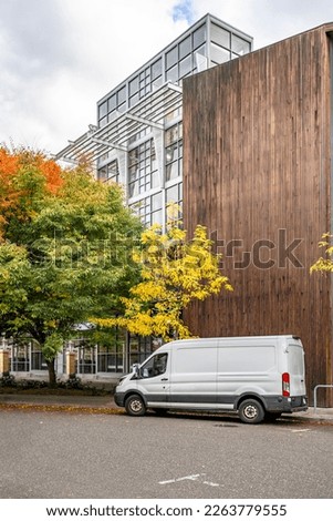 Commercial compact cargo small size white mini van delivered goods to client at multi-apartment high-rise building with wood wall and standing on the autumn street of urban city waiting for unloading