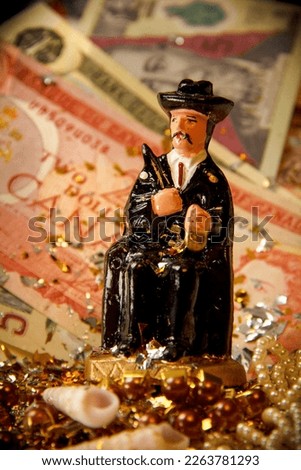 Close-up of the Figurine of a smoking man (Saint Simon from Guatemala) in front of international dollar bills, gold confetti and jewels, VERTICAL