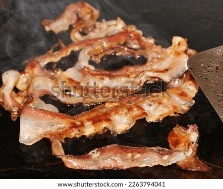 Iconic image for fast food. Strips of crispy bacon being cooked on the grill with a black background. Paddle for cooking. Food with runny fat. Real picture in the kitchen.