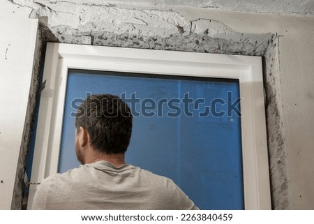 The proud homeowner, dressed in working clothes, as he installs a new energy-saving window in a window hole made in a concrete wall. The new window will help to reduce energy consumption.
