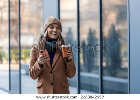 Cheerful young woman wearing coat walking outdoors, holding takeaway coffee cup, using mobile phone