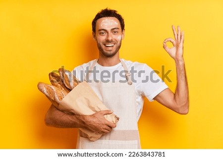 Indoor shot of cheerful caucasian man baker with satisfied expression makes ok gesture likes something smiles in agreement holds french baguettes in paper bag smiles widely wears apron isolated over Royalty-Free Stock Photo #2263843781