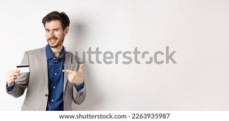 Successful male entrepreneur pointing at plastic credit card and smiling, recommending bank, standing on white background.