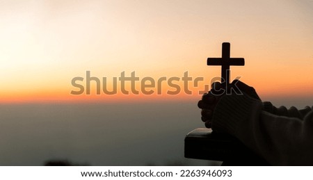 Silhouette of young woman kneeling down praying and holding christian cross for worshipping God at sunset background. concept of christian kneeling and praying to god.