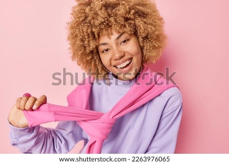 Happy curly haired woman smiles broadly shows white teeth ties jumper over shoulders feels glad looks directly at camera isolated over pink background. Positive emotions and feelings concept Royalty-Free Stock Photo #2263976065