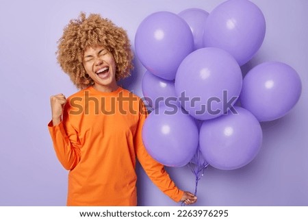 People special occasion and celebration concept. Optimistic woman wears orange jumper clenches fist celebrates special occasion holds bunch of inflated balloons isolated over purple background. Royalty-Free Stock Photo #2263976295
