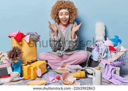 Surprised happy housewife wears headband and domestic robe spreads palms sits crossed legs on floor in disorganised room with different items around isolated over blue wall. Bring your house in order