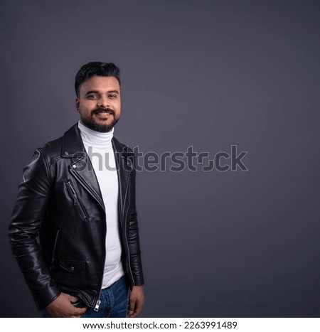 Young guy with dark hair mustache and beard wearing cool leather jacket and white jumper looking at camera and smiling.  square composition gray copy space. 