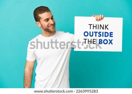 Handsome blonde man over isolated blue background holding a placard with text Think Outside The Box