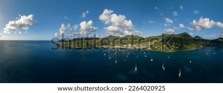 Panoramic picture of Anse d'arlet in Martinique, Caribbean island, with sailboats, turquoise water and cloudy blue sky in background.
