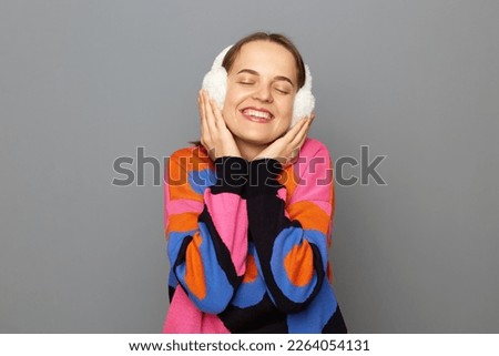 Portrait of relaxed satisfied woman with brown hair wearing sweater standing isolated over gray background, keeps eyes closed, enjoying her warm earmuffs.