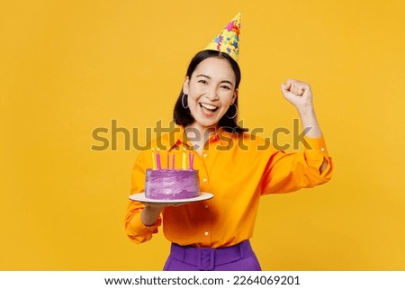 Happy fun overjoyed young woman wears casual clothes hat celebrating hold in hand purple cake with candles do winner gesture isolated on plain yellow background. Birthday 8 14 holiday party concept