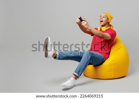 Full body young man of African American ethnicity he wear pink t-shirt yellow hat headphones sit in bag chair hold in hand play pc game with joystick console isolated on plain grey background studio