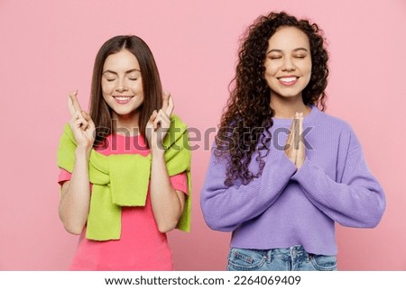 Young two friends women wears green purple shirts looking camera together wait for special moment, keep fingers crossed, hands folded in prayer gesture isolated on pastel plain light pink background Royalty-Free Stock Photo #2264069409