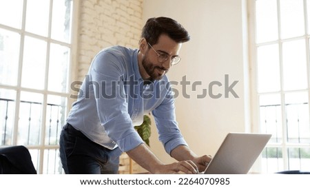 Happy young male manager employee in eyeglasses working on computer standing at table, typing email, communicating distantly with clients or colleagues or preparing electronic research report.
