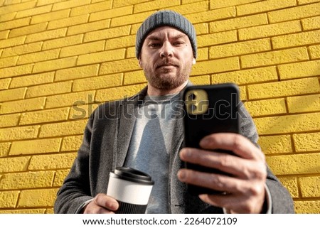 Adult man in hat and coat standing against yellow brick wall and taking picture with his smart phone.