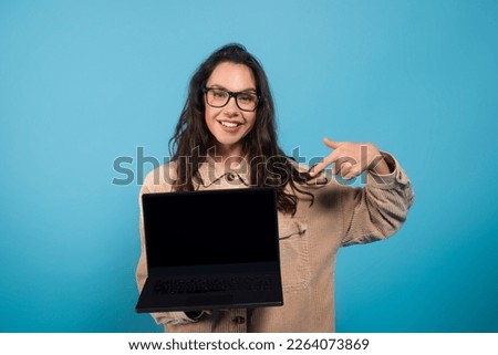 Cheerful millennial european female student in glasses point finger at laptop with empty screen, isolated on blue background, studio. Blogger advice, recommendation, website, study and work remotely