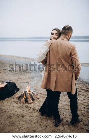a man and a woman stand on the shore of a lake near a fire. foggy cold weather. beige men's coat. bonfire on the shore of the reservoir. hug a loved one photo in beige tones. romantic photo