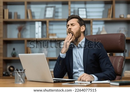 Mature man yawning inside classic office, senior businessman in workplace sleepless and overworked, boss in business suit sitting at desk, using laptop at work. Royalty-Free Stock Photo #2264077515