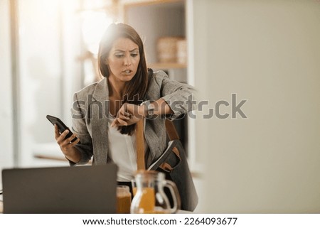 Shot of a multi-tasking young business woman looking on watch and using smartphone in her kitchen while getting ready to go to work. Royalty-Free Stock Photo #2264093677