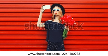 Portrait of beautiful young woman taking selfie with smartphone holding bouquet of red rose flowers in black round hat on background