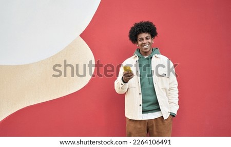 Happy young African American guy standing at red wall outdoors using cell phone, looking at camera holding cellphone enjoying doing online ecommerce shopping in mobile apps, chatting or playing games.