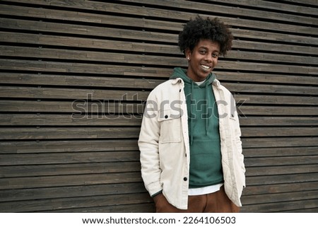 Young happy African American hipster teen guy standing at wooden wall background. Smiling cool stylish trendy gen z teenage student model looking at camera posing for authentic portrait outdoors. Royalty-Free Stock Photo #2264106503