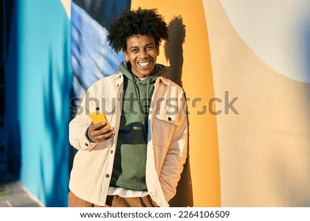 Smiling young cool African American guy holding mobile phone tech device standing at colorful city wall. Happy stylish authentic hipster teen boy using apps on cell outdoors, looking at camera. Royalty-Free Stock Photo #2264106509