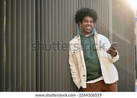 Cool smiling young African American guy holding mobile phone standing at wooden city wall. Happy stylish hipster teenager using cell outdoors, looking at camera with smartphone. Royalty-Free Stock Photo #2264106519
