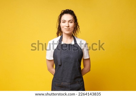 young girl waiter in uniform stands with her hands behind her back on a yellow background, a barista woman in a denim apron Royalty-Free Stock Photo #2264143835