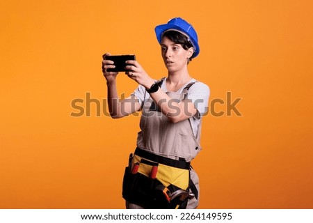 Construction worker taking pictures and photos with mobile phone, wearing hardhat as industrial builder in studio. Woman dressed as constructor capturing images on smartphone, photography.