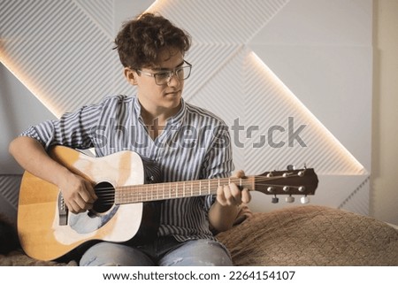 Modern man playing acoustic guitar learning musical performance sitting on couch at home. Teenager male casual guitarist chord bass solo melody instrumental music art hobby cultural entertainment Royalty-Free Stock Photo #2264154107
