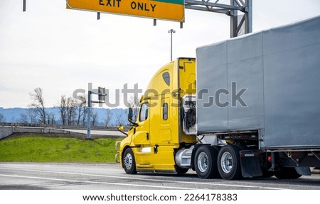 Bright yellow powerful long haul freight transportation big rig bonnet semi truck transporting goods in semi trailer covered with black rubberized fabric running on the wide highway with road signs Royalty-Free Stock Photo #2264178383