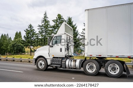 Big rig white industrial diesel day cab semi truck with roof spoiler and dry van semi trailer transporting commercial cargo load driving on the wide multiline highway road with trees on the side Royalty-Free Stock Photo #2264178427
