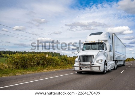 Powerful big rig white industrial grade high cab diesel semi truck with pipe grille guard transporting commercial cargo in dry van semi trailer running on the highway road with green hill Royalty-Free Stock Photo #2264178499