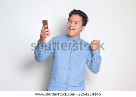 Portrait of Asian man in blue shirt raising his fist, celebrating success, winning game on his mobile phone. Isolated image on white background