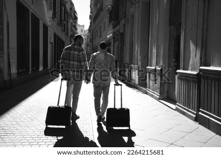 A young gay male couple walks down a street with their suitcases. The couple goes on a trip. The photo is taken from behind and in black and white. Vacation and travel concept. Royalty-Free Stock Photo #2264215681