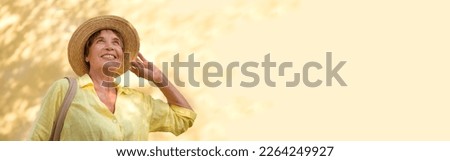 happy senior woman in sun hat walks  on summer city against the background of a yellow wall with shadows from trees. banner Royalty-Free Stock Photo #2264249927