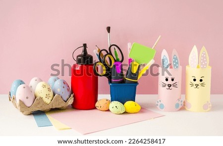Cute, funny rabbits made of colored paper on a pink background.  Craft for the Easter holiday, Easter eggs and office supplies are on the table.  Easter bright holiday concept.  Front view.