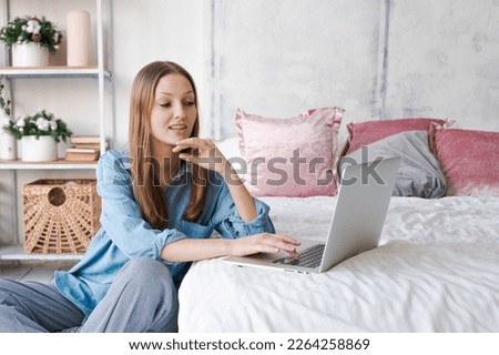 Young woman relaxes using laptop on cold winter day in bedroom while sitting on the floor by the bed. Woman checking social apps and work concept. Management and technology.